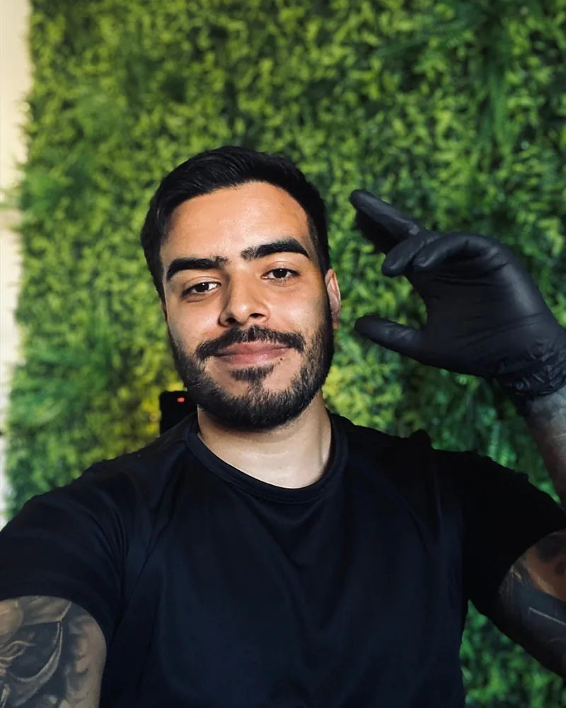 Junior sorrindo no estúdio, de camiseta preta e luva preta, em frente a uma parede de plantas
